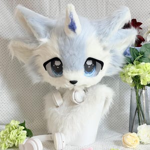 Peut inclure: Un masque de tête d'animal en peluche avec de la fausse fourrure blanche et bleu clair, une corne unique sur le front et de grands yeux bleus expressifs aux contours noirs. Le masque est présenté sur un support blanc avec un casque audio blanc.