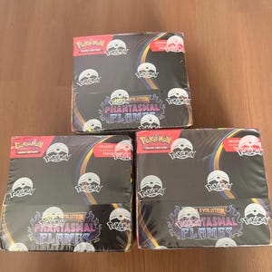 Puede incluir: Tres cajas selladas de cartas coleccionables Pokémon. Cada caja es negra con el texto "Pokémon" y "Evolution Phantasmal Flames" en blanco, morado y amarillo. Las cajas tienen un diseño de Poké Ball rojo y blanco.