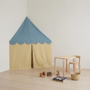 Può includere: Una tenda da gioco per bambini con tetto blu e pareti a righe gialle. Un tavolino e una sedia in legno sono nelle vicinanze, insieme ad animali giocattolo, un cestino e un libro. La tenda è allestita in un angolo di una stanza con pavimento in legno chiaro.
