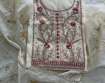 May include: An off-white garment featuring intricate silver embroidery, sequins, and beads. The design includes floral motifs with pink and orange accents, a central vertical beaded panel, and a decorative border along the hem and neckline.