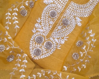 May include: A vibrant mustard yellow fabric featuring intricate white thread embroidery in a paisley-inspired pattern around the neckline. The design includes delicate leaf motifs and circular metallic embellishments scattered across the surface.