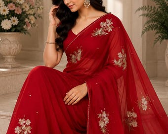 May include: A vibrant red saree with intricate floral embroidery in gold and white. The sheer fabric drapes elegantly, showcasing the delicate detailing. The saree features a V-neck design and is adorned with small, scattered embellishments.