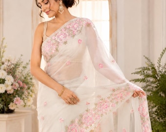 May include: A white saree with intricate pink floral embroidery along the borders and scattered across the sheer fabric. The saree is draped elegantly, showcasing the delicate design and texture. The model is wearing a matching blouse.