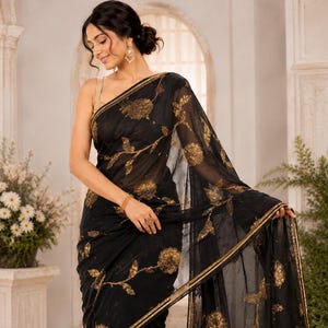 May include: Black sari with gold floral embroidery. The sheer fabric drapes elegantly, showcasing intricate golden flower and vine patterns. The sari features a gold border and is worn by a person.