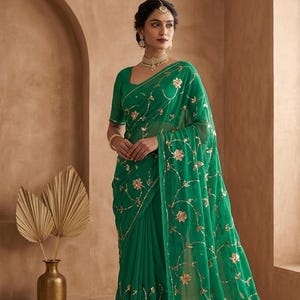 May include: A vibrant emerald green saree adorned with gold floral embroidery. The saree is worn with a matching blouse. The background features a neutral-toned wall and a decorative vase with dried palm leaves.