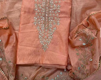 May include: A peach-colored fabric garment featuring intricate embroidery and reflective mirror accents. The design includes a central floral pattern around the neckline and decorative leaf-shaped mirror embellishments along the edges of the fabric.