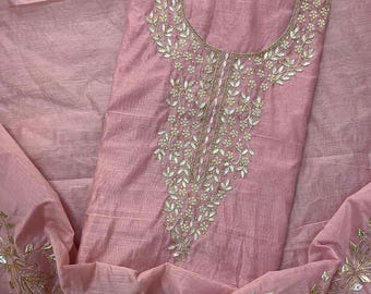 May include: A light pink fabric garment featuring intricate embroidery around the neckline and along the hem. The design includes metallic gold thread work and small, reflective, diamond-shaped mirror accents arranged in a floral vine pattern.