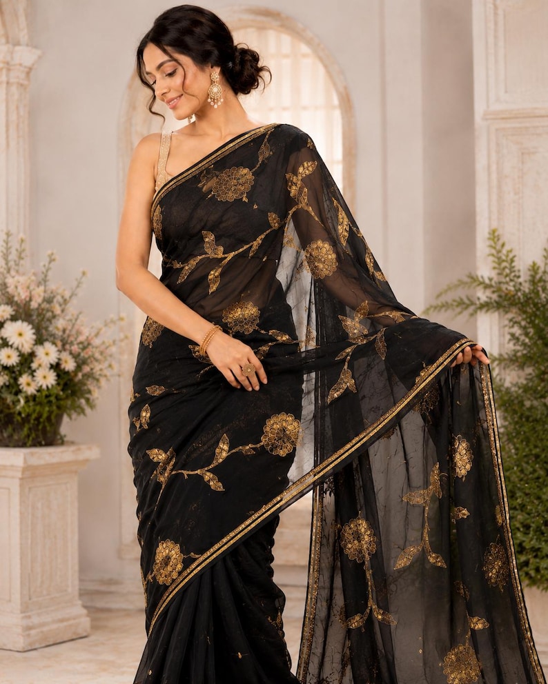 May include: Black sari with gold floral embroidery. The sheer fabric drapes elegantly, showcasing intricate golden flower and vine patterns. The sari features a gold border and is worn by a person.
