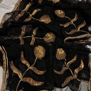 May include: Black sheer fabric with gold floral embroidery. The design features large, stylized flowers and leaves, with gold trim along the edges. The embroidery is detailed and adds a touch of elegance to the fabric.