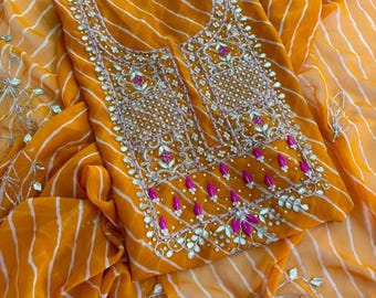 May include: A vibrant mustard yellow fabric featuring a white diagonal stripe pattern. The garment displays an intricate rectangular neckline panel adorned with silver metallic embroidery, reflective mirror-like accents, and small bright pink floral details.