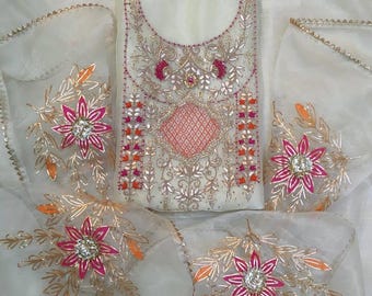 May include: A white sheer fabric garment featuring intricate embroidery with metallic gold thread, pink floral motifs, and orange accents. The design includes a central geometric pattern and scattered flower embellishments with sparkling centers.