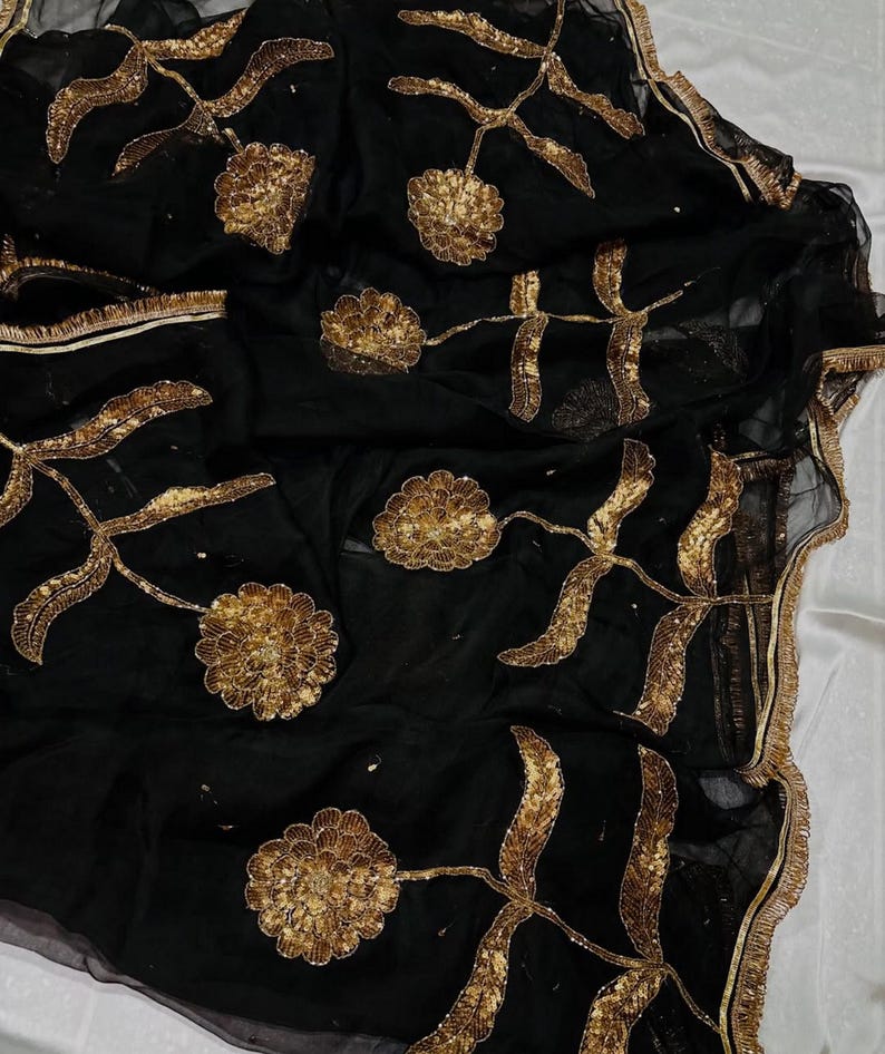 May include: Black sheer fabric with gold floral embroidery. The design features large, stylized flowers and leaves, with intricate detailing. The edges are trimmed with a gold border. The fabric is draped, creating soft folds and shadows.