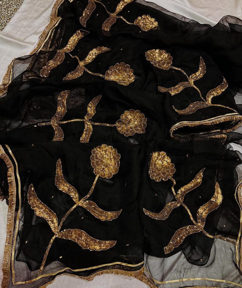 May include: Black sheer fabric with gold floral embroidery. The design features large, stylized flowers and leaves, with gold trim along the edges. The embroidery is detailed and adds a touch of elegance to the fabric.
