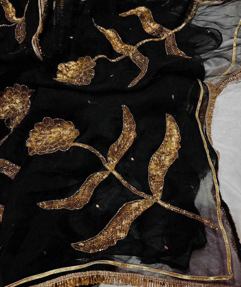 May include: Black fabric with gold floral embroidery. The design features large, stylized flowers and leaves with intricate sequin detailing. The edges are trimmed with a gold fringe.