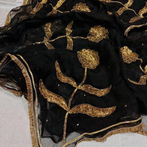 May include: Black sheer fabric with gold floral embroidery. The design features large, stylized flowers and leaves with intricate detailing. The edges are trimmed with a gold border and fringe, creating an elegant and ornate look.