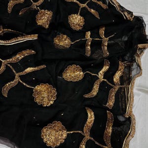 May include: Black sheer fabric with gold floral embroidery. The design features large, stylized flowers and leaves, with intricate detailing. The edges are trimmed with a gold border, adding a touch of elegance. The fabric is draped, creating soft folds and shadows.