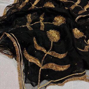 May include: Black sheer fabric with gold floral embroidery. The design features large, stylized flowers and leaves with intricate detailing. The edges are trimmed with a gold border and fringe, creating an elegant and ornate look.