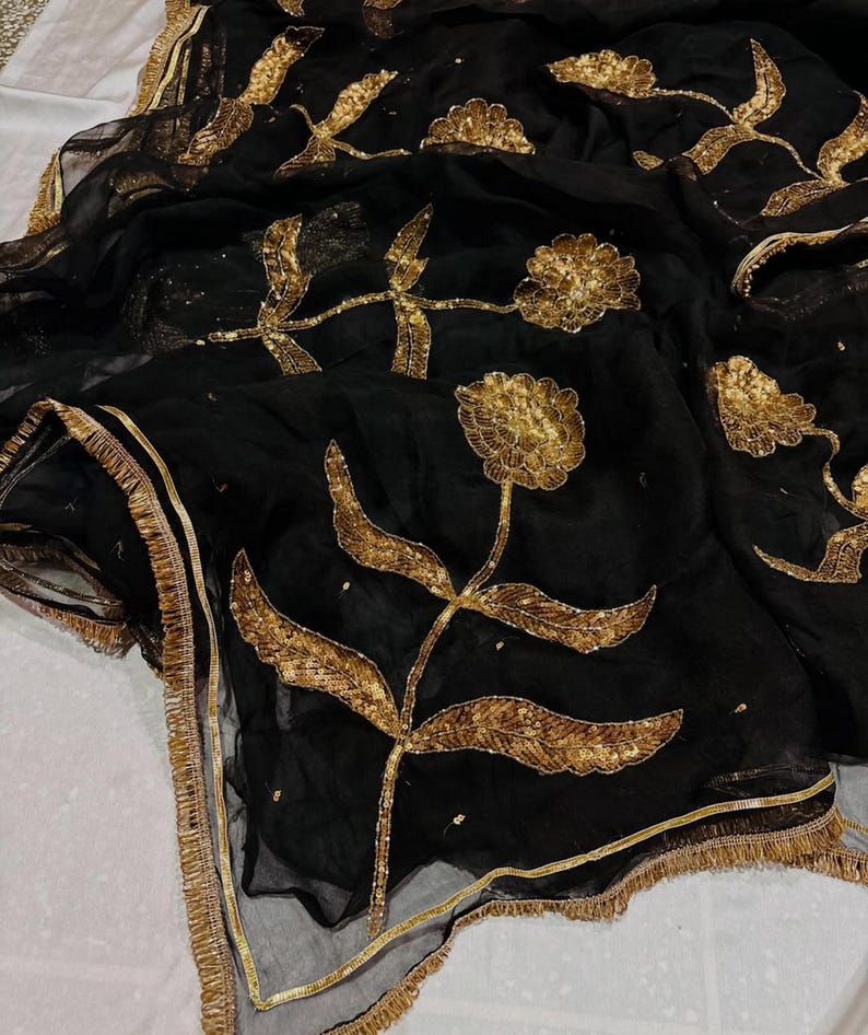 May include: Black sheer fabric with gold floral embroidery. The design features large, stylized flowers and leaves with intricate detailing. The edges are trimmed with a gold border and fringe, creating an elegant and ornate look.