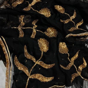 May include: Black fabric with gold floral embroidery. The design features detailed gold flowers and leaves, with gold trim along the edges. The fabric appears to be sheer, with a delicate texture.