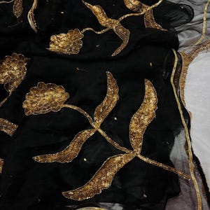 May include: Black fabric with gold floral embroidery. The design features large, stylized flowers and leaves with intricate sequin detailing. The edges are trimmed with a gold fringe.