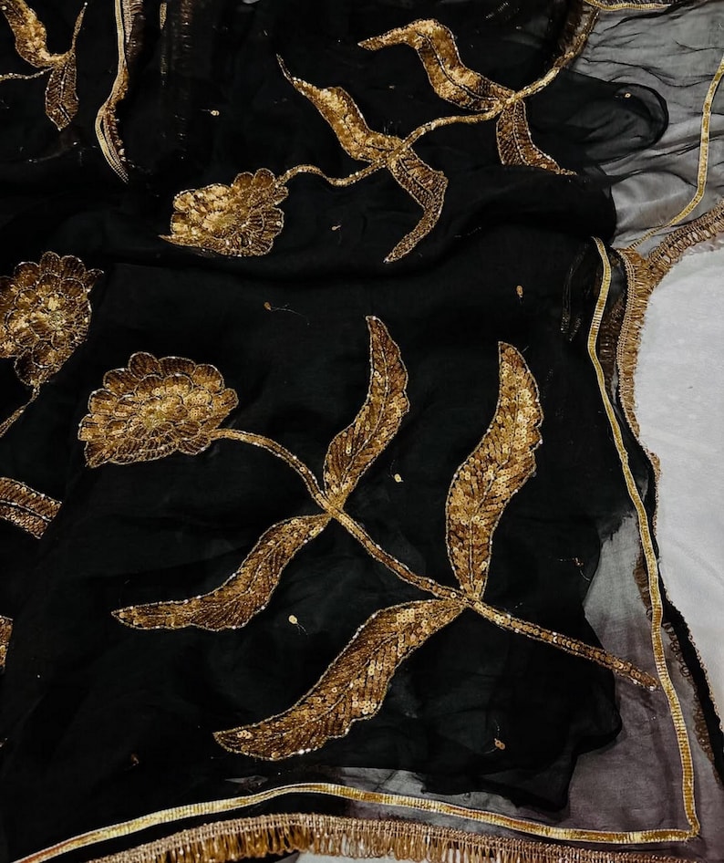 May include: Black fabric with gold floral embroidery. The design features large, stylized flowers and leaves with intricate sequin detailing. The edges are trimmed with a gold fringe.
