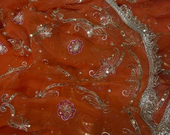 May include: An orange sheer fabric with intricate gold and silver embroidery. The design features floral and butterfly motifs, embellished with sequins. The edges are finished with a decorative border, creating a luxurious and ornate appearance.