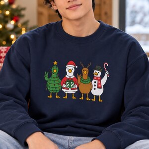 May include: A navy blue crewneck sweatshirt featuring a graphic of four ducks dressed for the holidays. One duck is a Christmas tree, one wears a Santa suit with a gift, one is a reindeer with a wreath, and one is a snowman with a candy cane.
