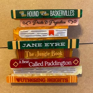 May include: A decorative stack of books featuring titles including The Hound of the Baskervilles, Pride & Prejudice, Jane Eyre, The Jungle Book, A Bear Called Paddington, and Wuthering Heights. The stack is made of colourful resin with varied textures.