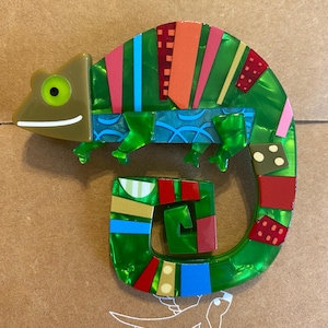 May include: A brooch shaped like a chameleon, featuring a glossy green body with a brown head and a bright green eye. The body is decorated with colourful geometric patterns in red, blue, pink, and gold, and the tail is curled into a loop.