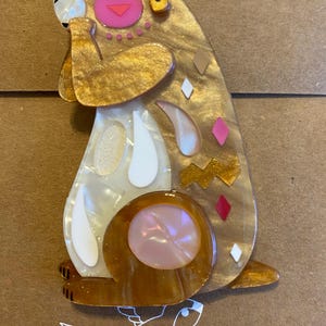 May include: A brooch shaped like a sitting rabbit, crafted from layered acrylic in shades of gold, brown, and pearlescent white. The design features a pink circular cheek, a small triangular nose, and decorative geometric accents in pink and white.