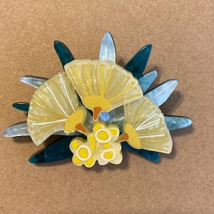 May include: A floral brooch with three yellow fan-shaped elements with white detailing, accented by yellow and orange centres. The design includes teal and light blue leaf-shaped elements, creating a layered, decorative effect.