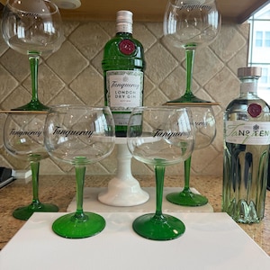 Op de afbeelding: Een verzameling heldere wijnglazen met groene stelen en voetjes, samen met twee gin-flessen. De glazen zijn voorzien van het opschrift "Tanqueray". Eén fles is groen met een rode zegel, de andere is helder.