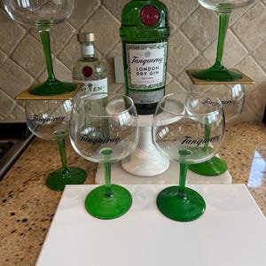 Może przedstawiać: Zestaw kieliszk&oacute;w do ginu z zielonymi n&oacute;żkami i napisem "Tanqueray". Widoczna jest r&oacute;wnież zielona butelka Tanqueray London Dry Gin oraz mniejsza butelka ginu No. TEN. Kieliszki ustawione są na białej powierzchni.