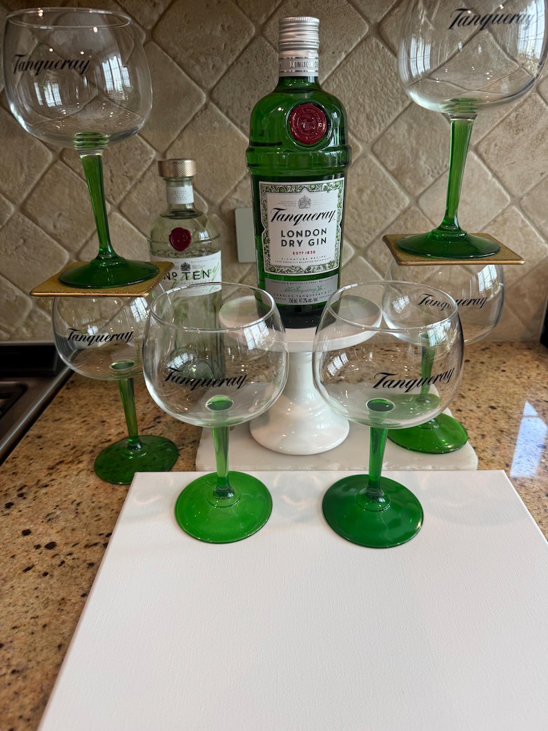 Może przedstawiać: Zestaw kieliszk&oacute;w do ginu z zielonymi n&oacute;żkami i napisem "Tanqueray". Widoczna jest r&oacute;wnież zielona butelka Tanqueray London Dry Gin oraz mniejsza butelka ginu No. TEN. Kieliszki ustawione są na białej powierzchni.