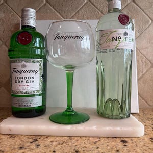Może przedstawiać: Dwie zielone butelki ginu i duży kieliszek z napisem "Tanqueray". Butelki są oznaczone jako "Tanqueray London Dry Gin" i "Tanqueray No. TEN". Kieliszek ma zieloną n&oacute;żkę i podstawę. Na białej marmurowej tacy.