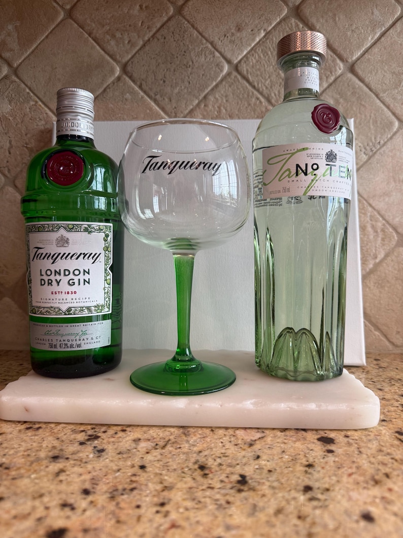 Może przedstawiać: Dwie zielone butelki ginu i duży kieliszek z napisem "Tanqueray". Butelki są oznaczone jako "Tanqueray London Dry Gin" i "Tanqueray No. TEN". Kieliszek ma zieloną n&oacute;żkę i podstawę. Na białej marmurowej tacy.