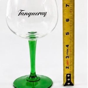 Może przedstawiać: Przezroczysty kieliszek do ginu z zieloną n&oacute;żką i podstawą, z napisem "Tanqueray" czarną czcionką. Kieliszek ma około 18 cm wysokości, obok ż&oacute;łtej miarki.