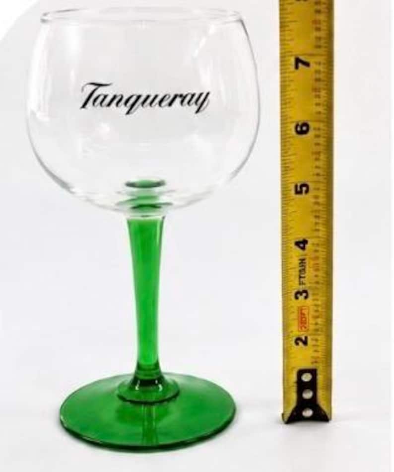 Może przedstawiać: Przezroczysty kieliszek do ginu z zieloną n&oacute;żką i podstawą, z napisem "Tanqueray" czarną czcionką. Kieliszek ma około 18 cm wysokości, obok ż&oacute;łtej miarki.