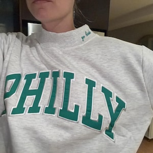 May include: A light gray heathered mock neck sweatshirt featuring the word PHILLY in large, bold green letters with a white outline across the chest. The collar includes the embroidered text go birds in a small, cursive green font.