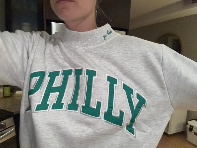 May include: A light gray heathered mock neck sweatshirt featuring the word PHILLY in large, bold green letters with a white outline across the chest. The collar includes the embroidered text go birds in a small, cursive green font.