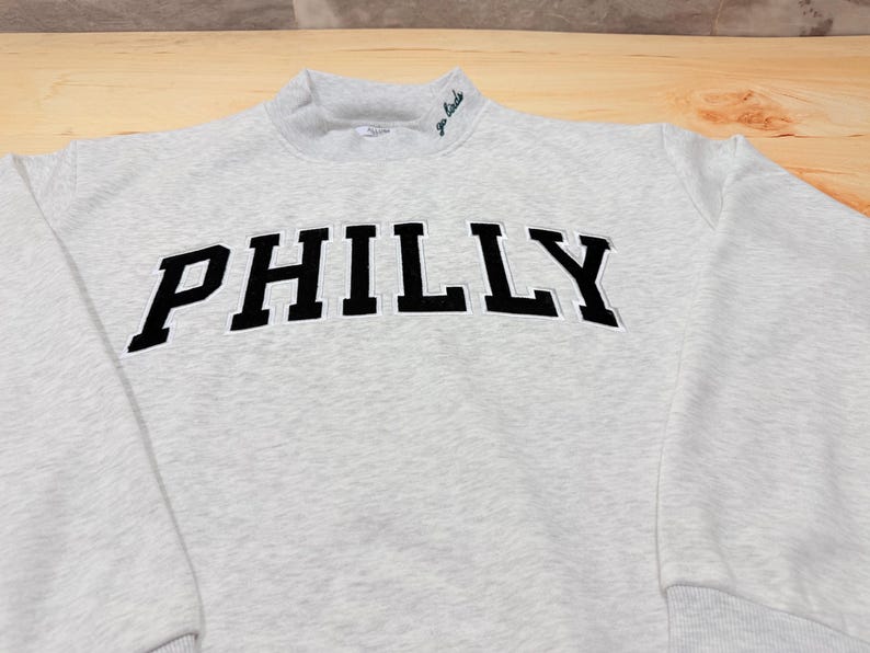 May include: A light gray heathered sweatshirt featuring the word PHILLY in large black block letters across the chest. The garment has a mock neck collar with the small embroidered text go birds in dark green on the right side of the neckline.