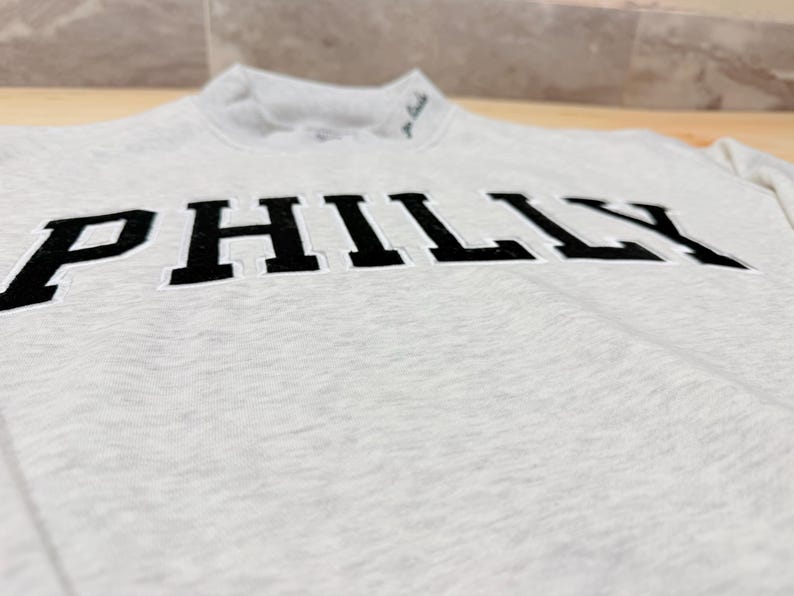 May include: A light gray heathered sweatshirt featuring the word PHILLY in large, bold, black block letters across the chest. The garment has a mock neck collar with small, dark embroidered text visible near the neckline on the left side.