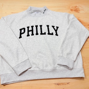 May include: A light gray heathered sweatshirt featuring the word PHILLY in bold black block letters across the chest. The garment has a mock neck collar with the text go birds embroidered in small black script on the right side of the neckline.