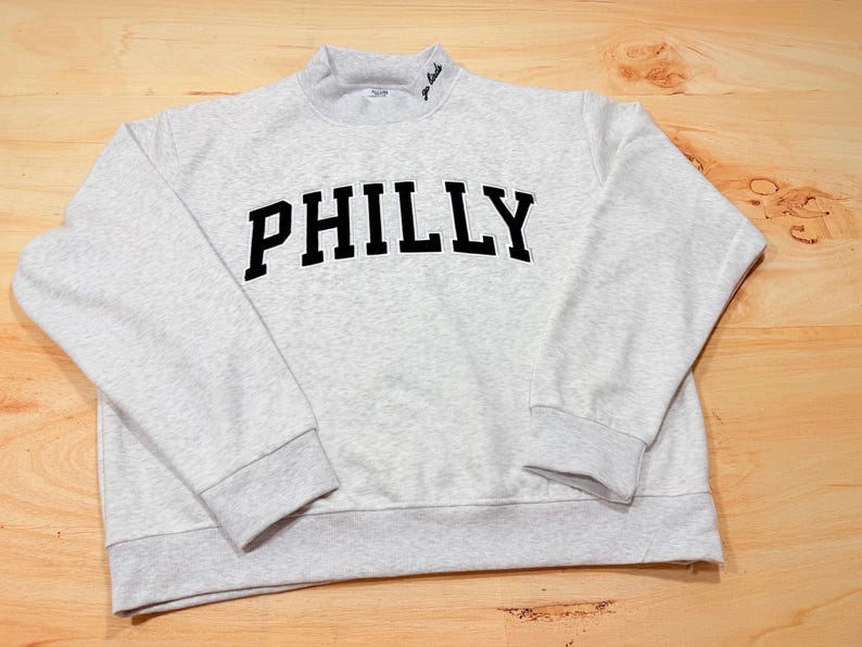 May include: A light gray heathered sweatshirt featuring the word PHILLY in bold black block letters across the chest. The garment has a mock neck collar with the text go birds embroidered in small black script on the right side of the neckline.