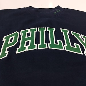 May include: A navy blue crewneck sweatshirt featuring the word PHILLY in large, arched green letters with a white outline. The text go birds is embroidered in small green cursive script on the collar near the neckline of the garment.