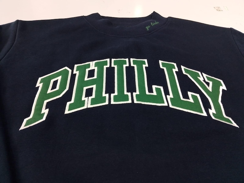 May include: A navy blue crewneck sweatshirt featuring the word PHILLY in large, arched green letters with a white outline. The text go birds is embroidered in small green cursive script on the collar near the neckline of the garment.