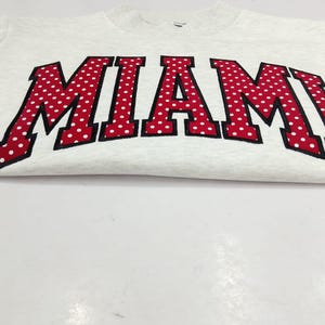 May include: A light gray heathered crewneck sweatshirt featuring the word MIAMI in large, arched, red block letters with a white polka dot pattern and a thick black outline. The text is centered across the chest of the garment.