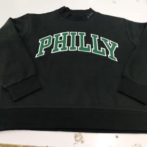 May include: A black long-sleeve sweatshirt featuring the word PHILLY printed in a large, arched, green font with a white outline across the chest. The garment has a crew neckline, ribbed cuffs, and a ribbed hemline in a matching black fabric.