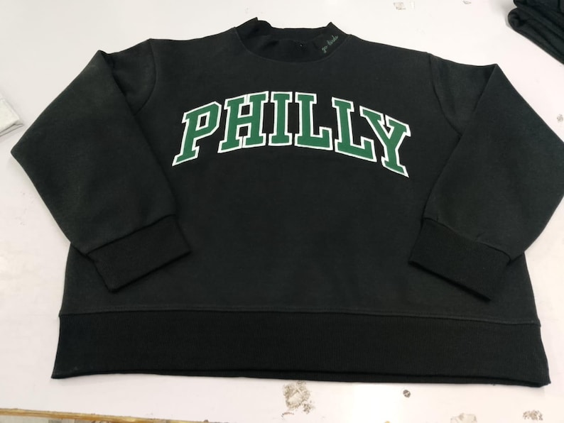 May include: A black long-sleeve sweatshirt featuring the word PHILLY printed in a large, arched, green font with a white outline across the chest. The garment has a crew neckline, ribbed cuffs, and a ribbed hemline in a matching black fabric.