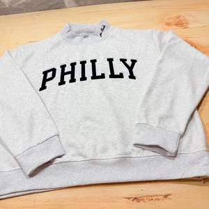 May include: A light gray heathered sweatshirt featuring the word PHILLY in bold black block letters across the chest. The garment has a mock neck collar with small black embroidered text on the right side, long sleeves, and ribbed cuffs.
